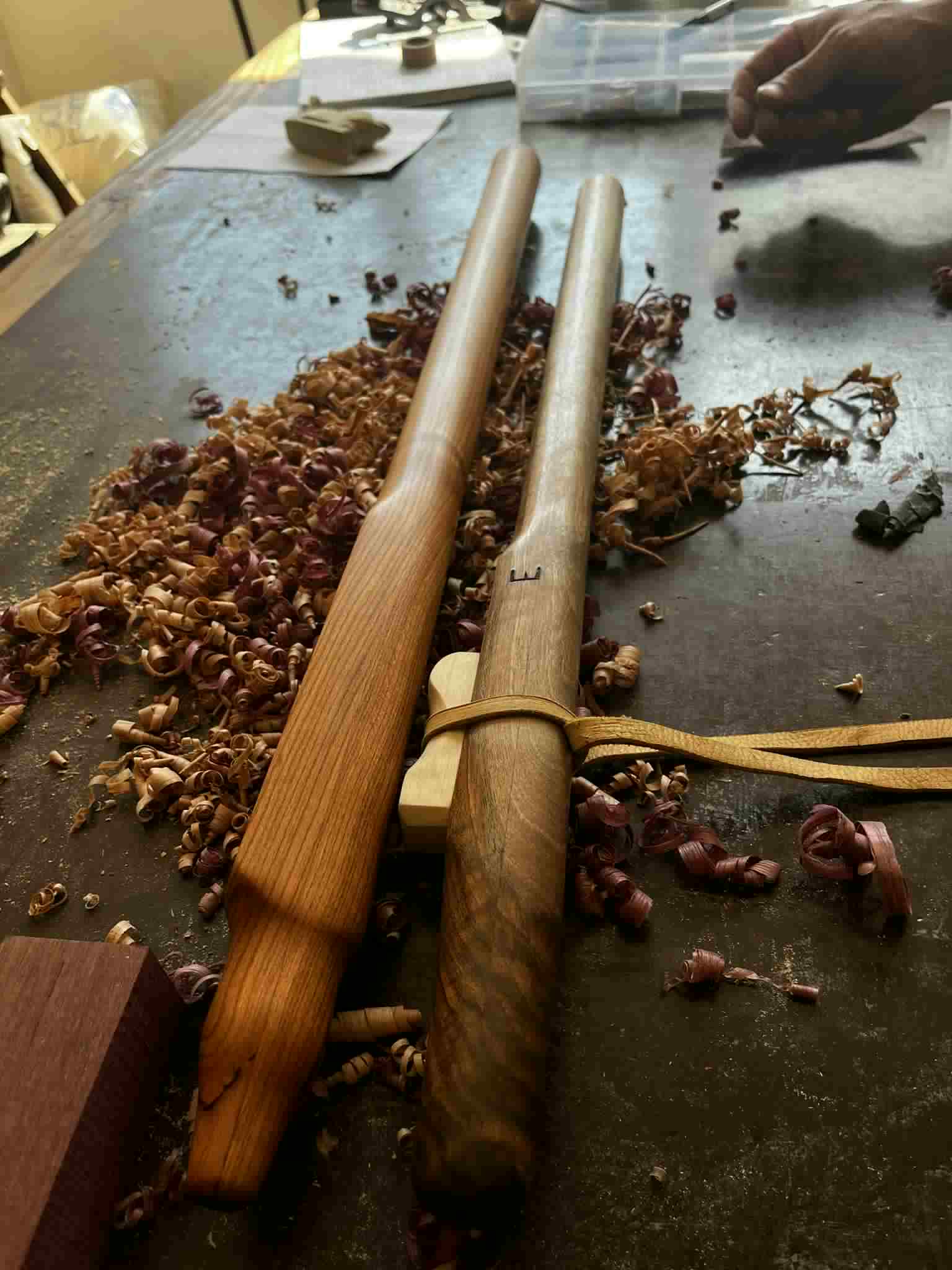 River Song and a second flute side by side on the bench, surrounded by fresh wood shavings as Radenko works